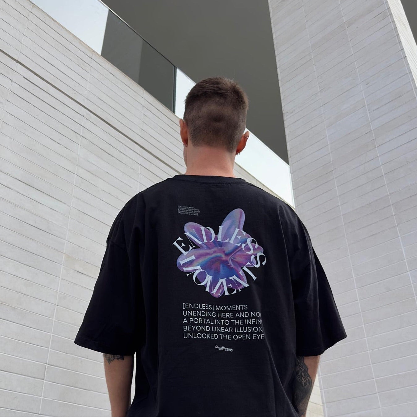 Model wearing black oversized t-shirt: the luxury streetwear 'Endless Moments' graphic tee with a pink and purple futuristic shape, white text paragraph beneath it. Drop shoulder tee, model facing urban wall and pillar