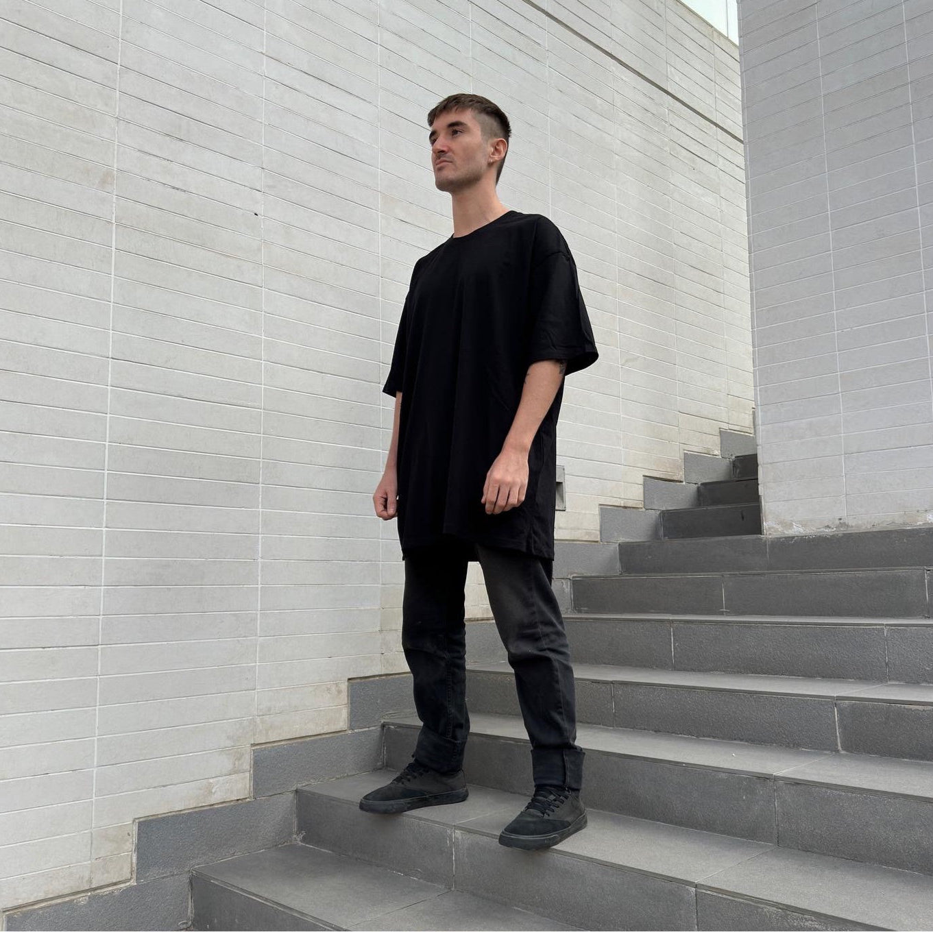 Model wearing full black streetwear fit standing on grey staircase, bottom diagonal perspective