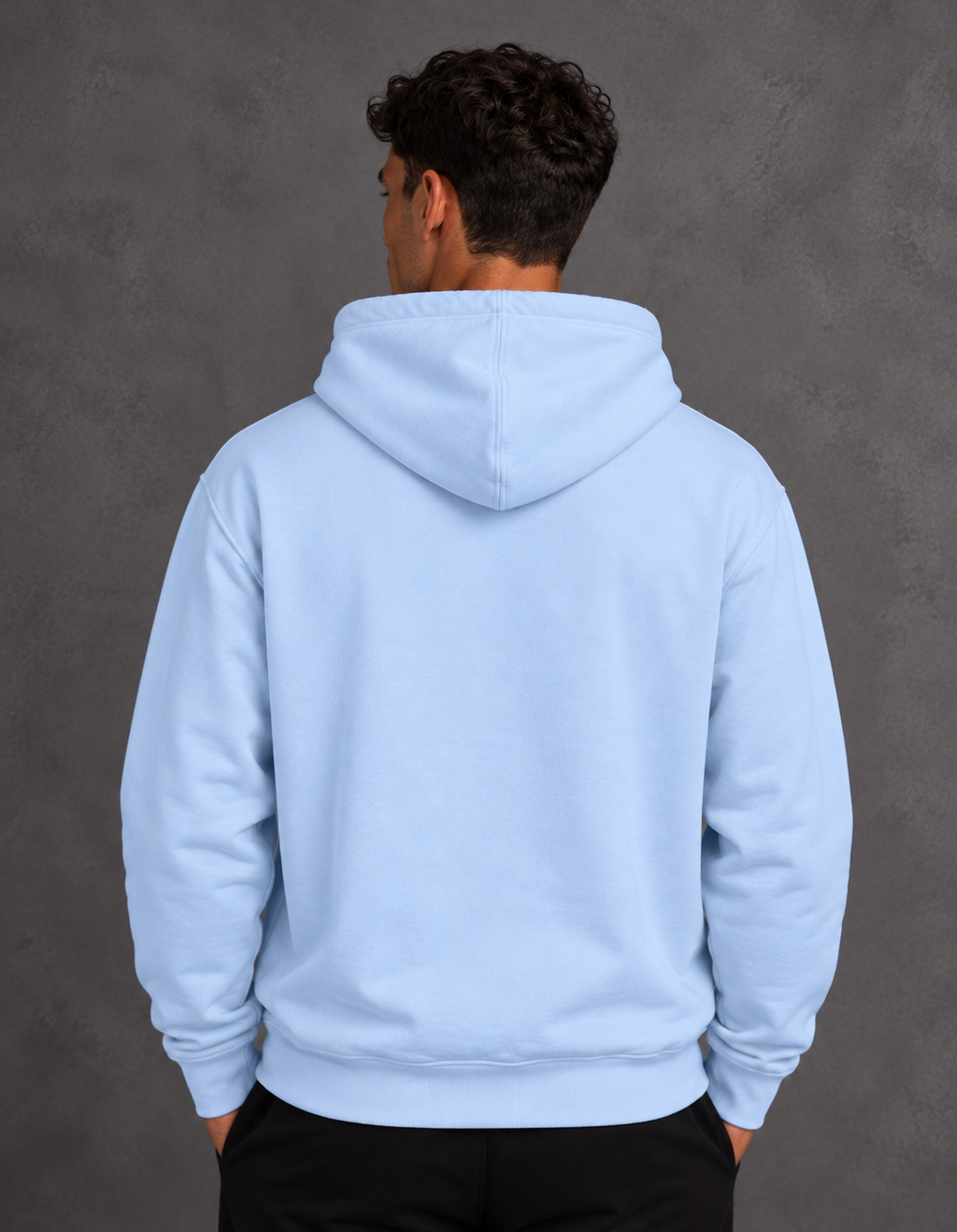 Back view of a model wearing a plain light blue hoodie with the hood lowered, hands in the black pant's pockets.
