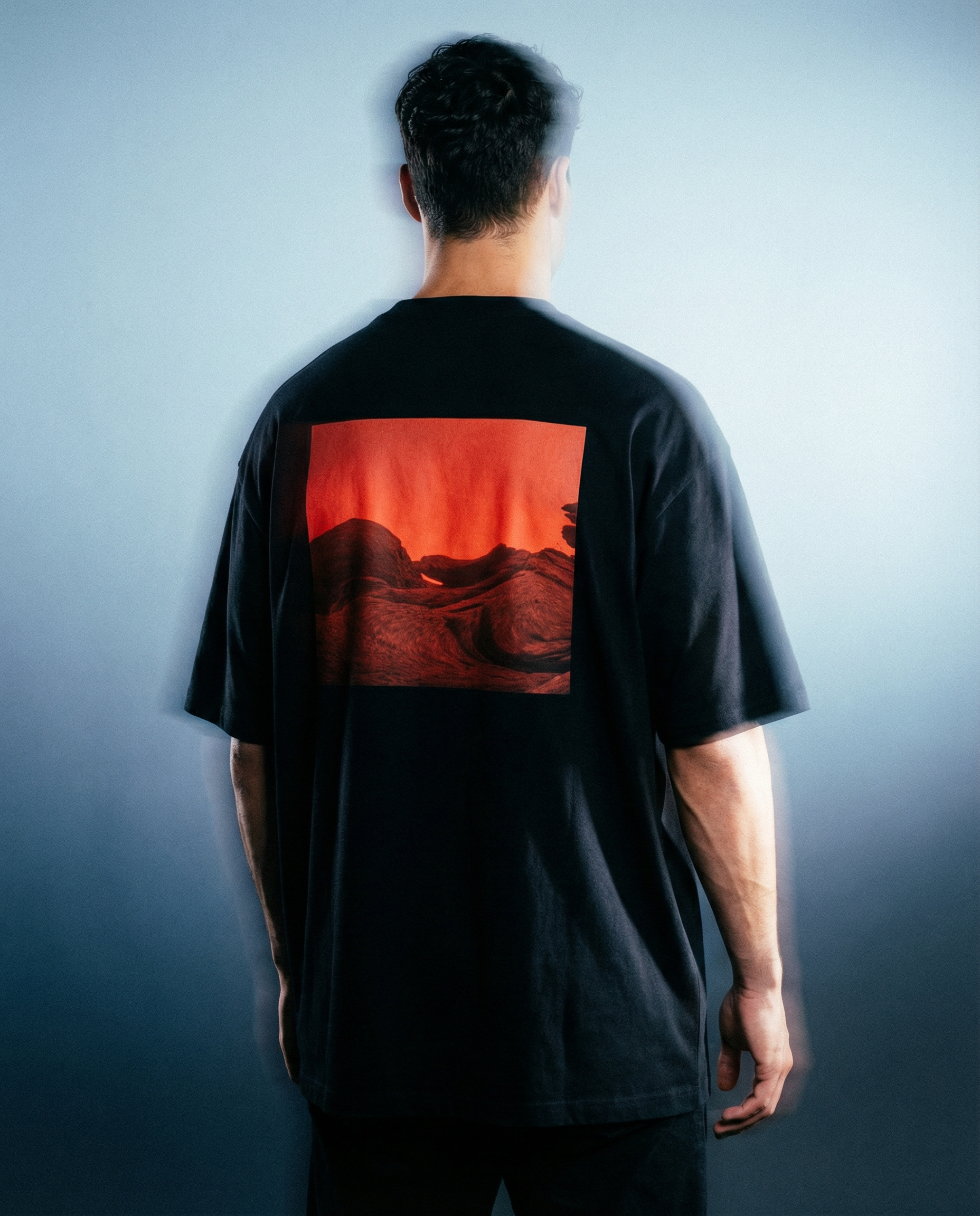 Person wearing an oversized black t-shirt with drop shoulder, red  graphic print on the back against a light blue gradient wall, in luxury streetwear urban clothing aesthetic