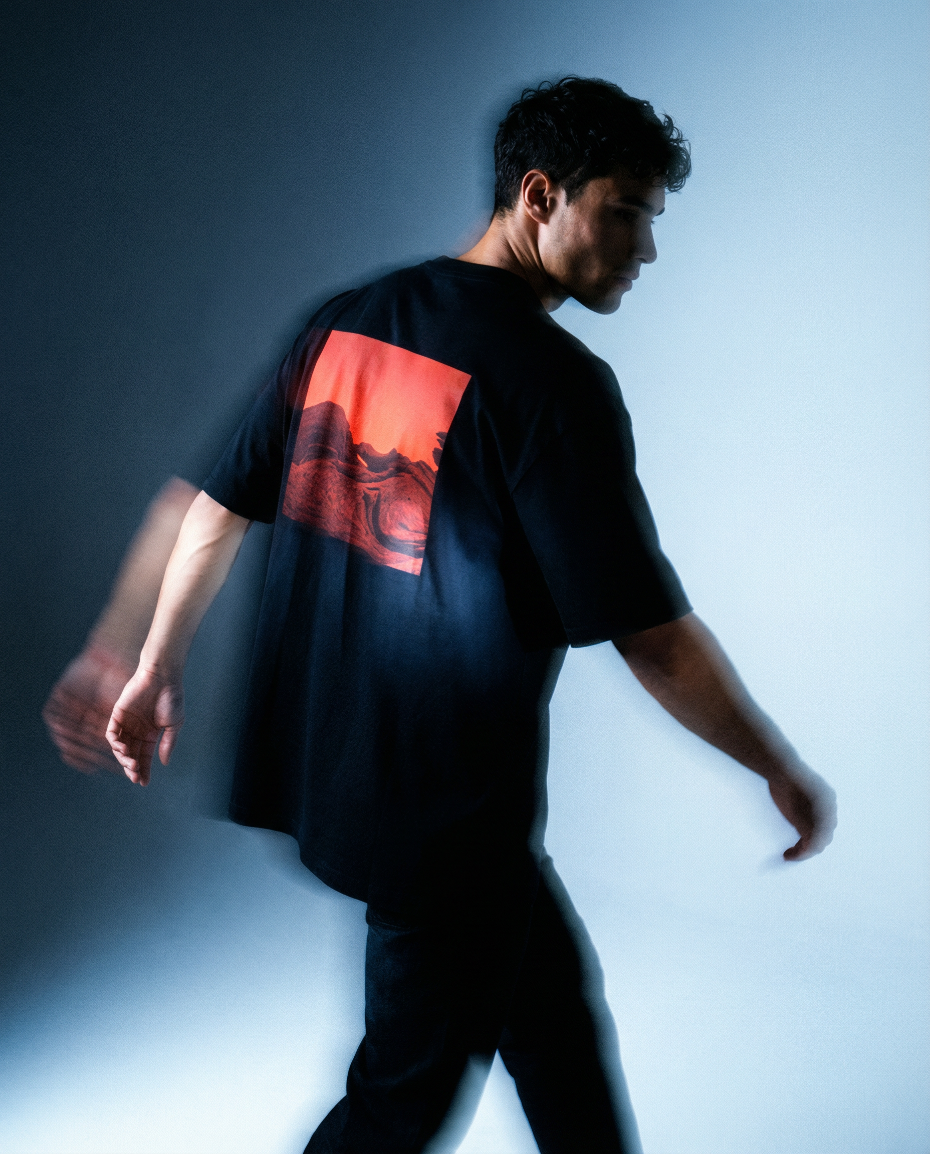 Person wearing an oversized black t-shirt with drop shoulder sleeves, red graphic back print, gradient background in luxury streetwear urban clothing aesthetic AETERIUS