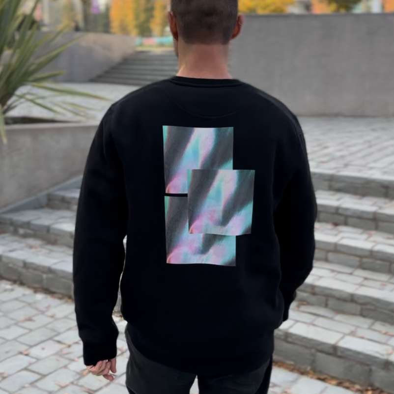 black crewneck sweatshirt with iridescent graphic on the back, model walking in urban area with staircase