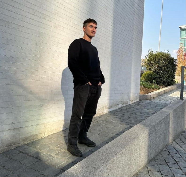 Luxury streetwear male model wearing black crewneck sweatshirt with hands in jeans pockets standing in urban sidewalk against grey wall