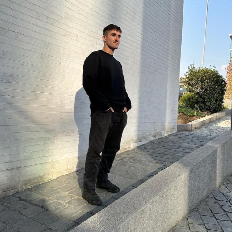 Luxury streetwear male model wearing black crewneck sweatshirt with hands in jeans pockets standing in urban sidewalk against grey wall