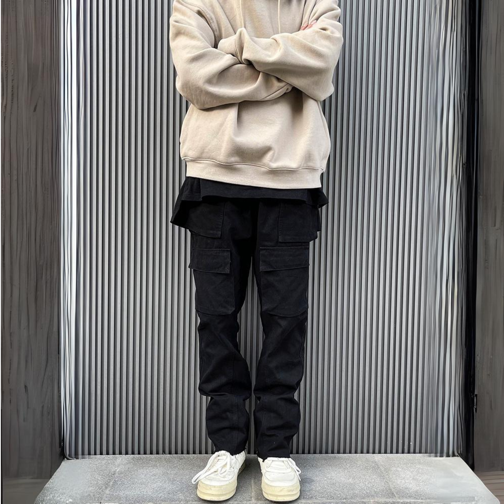 model in mens luxury streetwear fit of black cargo pants, beige sweatshirt and white shoes, standing against grey urban wall with stripes